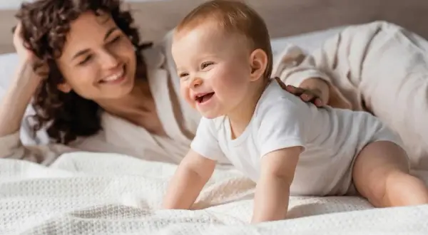 Un bebé de 7 meses gatea en la cama, riendo mientras su madre lo observa con una sonrisa.