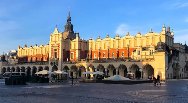 Sukiennice Kraków w blasku słońca, z arkadami i wieżą zegarową. Rynek tętni życiem.