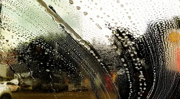 Tłusta szyba w samochodzie pokryta pianą i kroplami wody. Widok na rozmazane drzewa i światła uliczne.