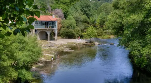 Disfrutando del sol y el agua en la playa fluvial Mondariz. Un edificio con arcos y tejado rojo se alza junto al río, donde unas personas se refrescan.