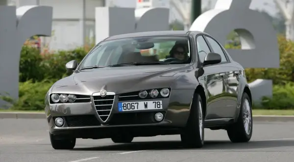 Eleganckie Alfa Romeo 159 2.4 JTDm w kolorze grafitowym, z kobietą za kierownicą, jedzie po drodze.