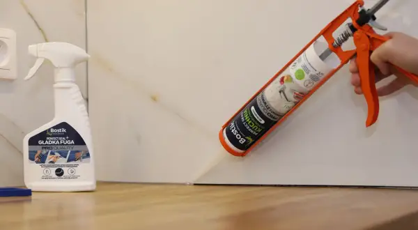 Aplikacja silikonu do blatu kuchennego. Pistolet z kartuszem Bostik Perfect Seal Kuchnia aplikuje masę uszczelniającą. Obok stoi butelka preparatu do wygładzania fug.
