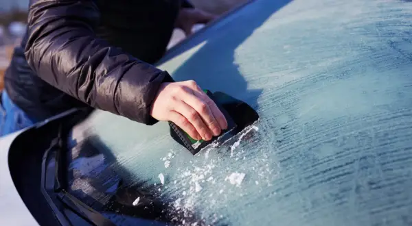 En hand skrapar is från insidan av bilrutan. Snöflingor syns på den frostiga ytan.