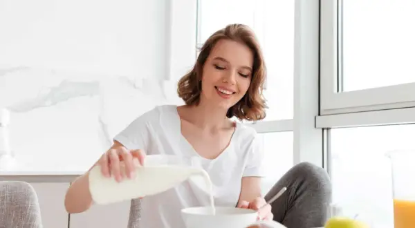 Uśmiechnięta dziewczyna nalewa mleko do miski, dbając o swoją masę kostną. Na stole croissant i jabłko.