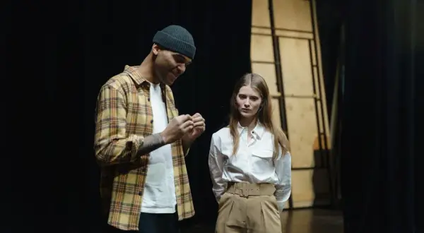 Mężczyzna w kratę i czapce rozmawia z kobietą na scenie. Czy to próba przed występem?
