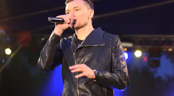 Artysta liber śpiewa do mikrofonu na scenie, ubrany w czarną kurtkę.