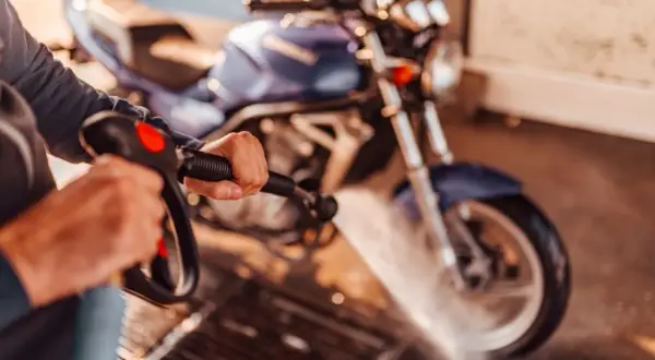 Persona limpiando una moto azul con una hidrolavadora. El agua salpica sobre la rueda delantera.