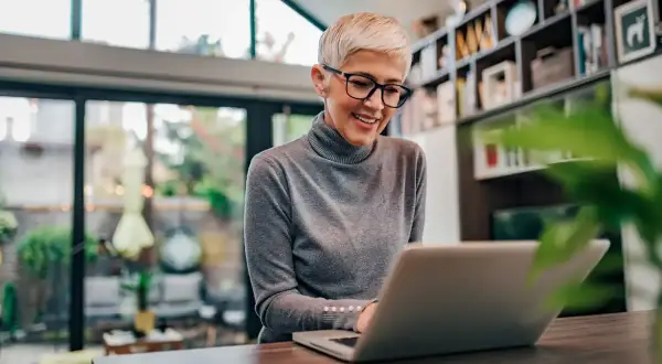Uśmiechnięta kobieta z krótkimi blond włosami i okularami pracuje na laptopie, szukając informacji, czy na umowę zlecenie należy się dodatek nocny.