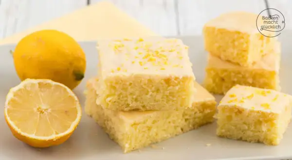 Saftiger Zitronenkuchen vom Blech, bestreut mit Zitronenschale. Daneben liegen frische Zitronen.