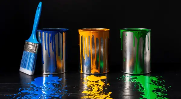 Tre barattoli di vernice (blu, giallo, verde) con gocce e schizzi su un tavolo scuro. Un pennello blu accanto. Il solvente cos'è?