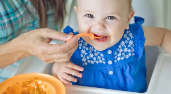 Ein Baby in einem blauen Kleid erhält Brei vom Löffel. Die Augen des Babys strahlen, ein Zeichen für die ersten reifezeichen baby.