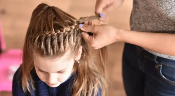 Bambina con capelli castani corti e trecce a cascata. Le mani di una persona le stanno creando una nuova treccia.