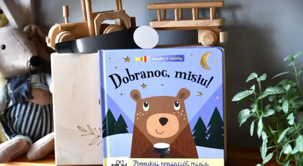 Książka "Dobranoc, misiu!" z latarką, obok pluszowego misia i drewnianych zabawek.
