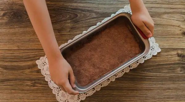 Mani che preparano un tiramisù senza caffè, con cacao in polvere in una teglia su un centrino di pizzo.