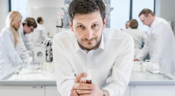 Mężczyzna w białej koszuli trzyma fiolkę z płynem, w tle laboratorium, gdzie pracują inni naukowcy. To kluczowe xtpl akcje.