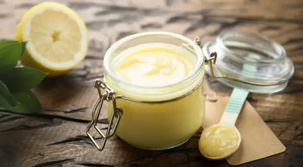 Un pot de lemon curd onctueux, une moitié de citron et des feuilles vertes sur fond de bois. Une cuillère en bois est prête à déguster.