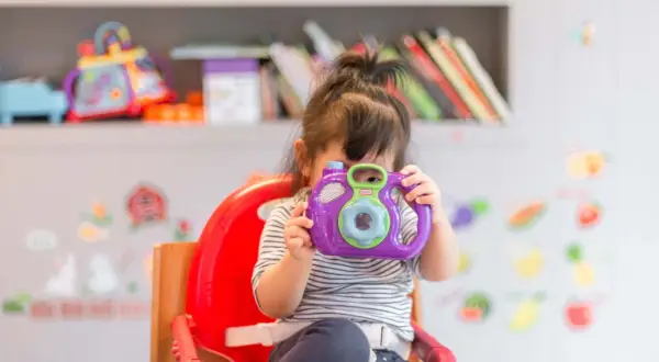 A young child in a striped shirt sits in a red high chair, holding a purple toy camera to their face.