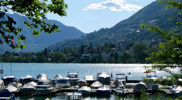 Båtar täckta med presenningar vid en brygga vid Lago Maggiore. Berg och en stad syns i bakgrunden.