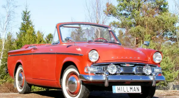 En röd cabriolet, en klassisk Hillman bil, står parkerad utomhus bland träd.