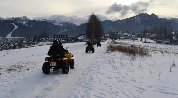 Ludzie na quadach jadą przez ośnieżony teren z widokiem na góry i zabudowania.