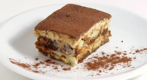 Une part de tiramisu, avec sa traduction française, est saupoudrée de cacao sur une assiette blanche.