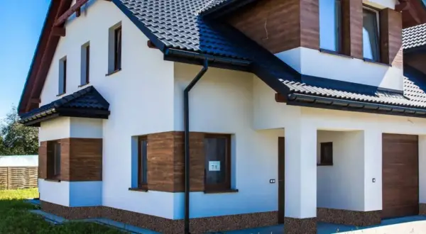 Nowy dom z białym tynkiem i drewnianymi akcentami. Brak cokołu na elewacji, widoczna kamyczkowa podmurówka.