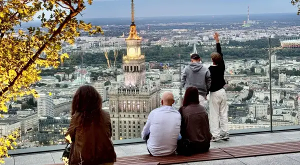 Ludzie podziwiają panoramę Warszawy z Pałacem Kultury i Nauki. Widać wiele warszawskich wież i budynków.