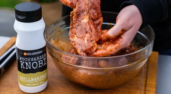 Fleisch wird mit einer würzigen Grillmarinade in einer Schüssel vermischt. Die Flasche "#Schwarzer Knobi" zeigt, wie einfach es ist, Grillmarinade selber machen.