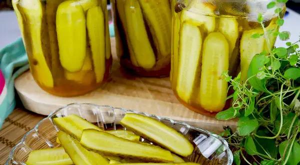 Słoiki pełne ogórków z curry, gotowe do słoików. Na talerzyku pokrojone ogórki, obok świeża zielenina.