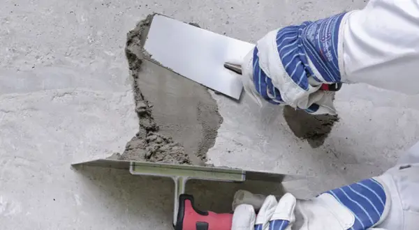 Naprawa betonu na zewnątrz. Ręce w rękawicach nakładają zaprawę na uszkodzony fragment ściany za pomocą kielni.
