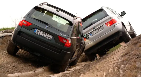 Dwa BMW E83, zielone i srebrne, pokonują trudny teren, wspinając się po piaszczystym zboczu z drewnianymi przeszkodami.