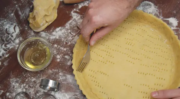 Mani che preparano una pasta frolla morbida, bucherellandola con una forchetta in una tortiera. Farina e ingredienti sparsi sul tavolo.