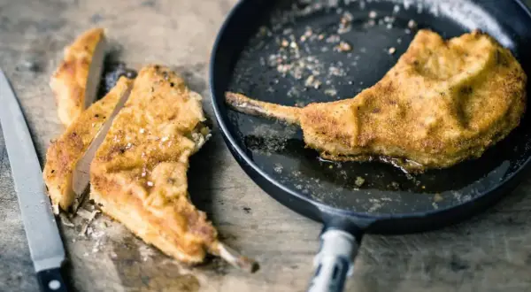 Ein paniertes Kotelett braten in der Pfanne. Daneben liegen bereits geschnittene Stücke auf einem Holzbrett, daneben ein Messer.