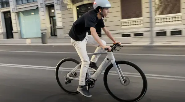 Un joven con casco y ropa casual pedalea una bicicleta eléctrica por la ciudad.