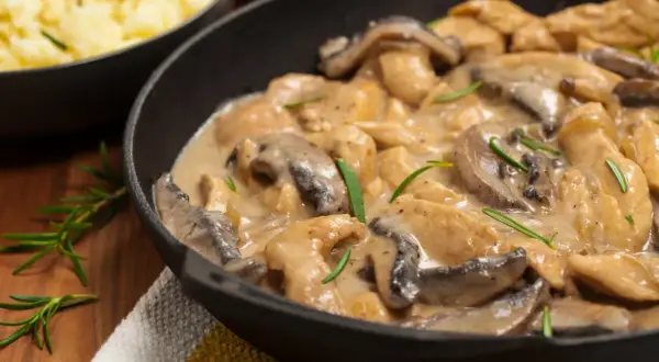 Pyszny kurczak w sosie pieczarkowym, jak u Ani gotuje, w czarnej patelni, z gałązkami rozmarynu. Obok miska z purée.