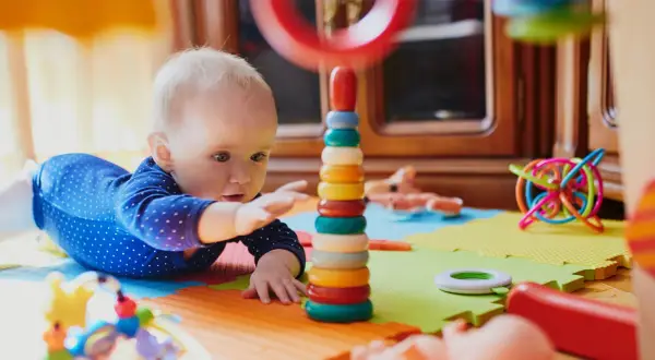 Zaintrygowany 6 miesięczne dziecko sięga do kolorowej wieży z kółek, otoczony zabawkami na macie.