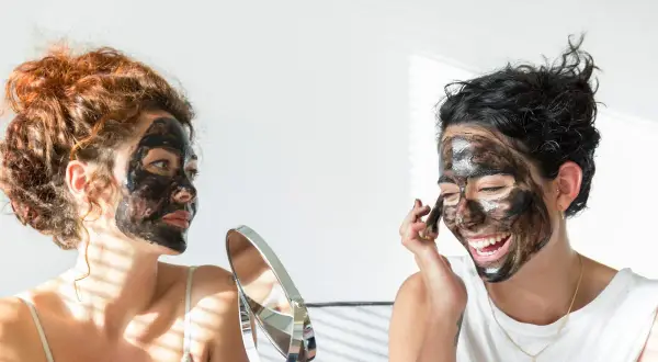 Dos mujeres con mascarilla negra en el rostro, una se mira en un espejo, buscando quitar espinillas negras grandes.
