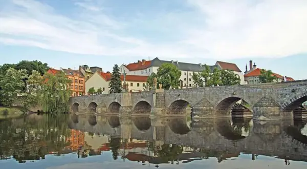 Historische Steinbrücke über den Fluss in Písek, Tschechien, mit Spiegelung im Wasser und bunten Häusern im Hintergrund.