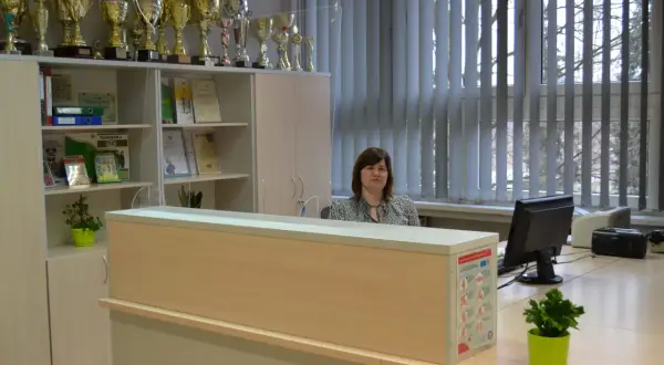 Kobieta siedzi za ladą w sekretariacie, wśród licznych pucharów i dyplomów.