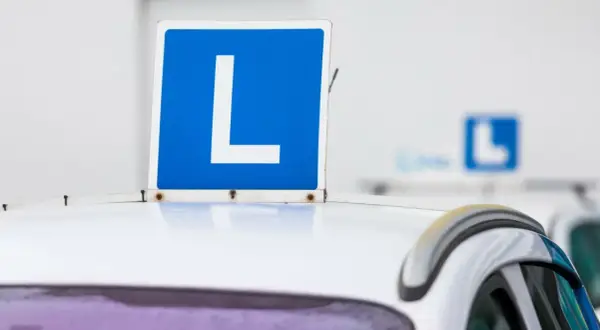 Tablica "L" na dachu białego samochodu. Zbliża się egzamin na prawo jazdy, a ten znak symbolizuje naukę jazdy.