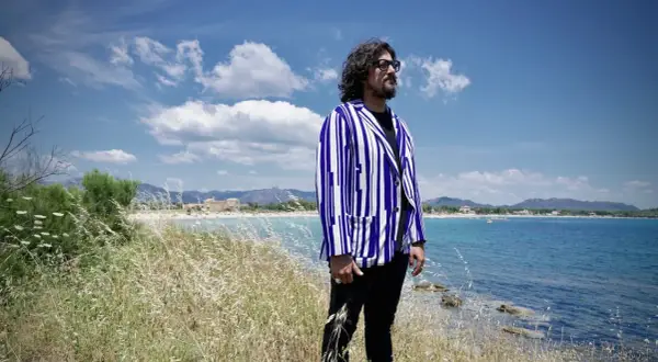 Uomo con giacca a righe blu e bianche guarda il mare in Sardegna, tra i campi di erba alta. Un'ambientazione perfetta per scoprire i **4 ristoranti Sardegna**.