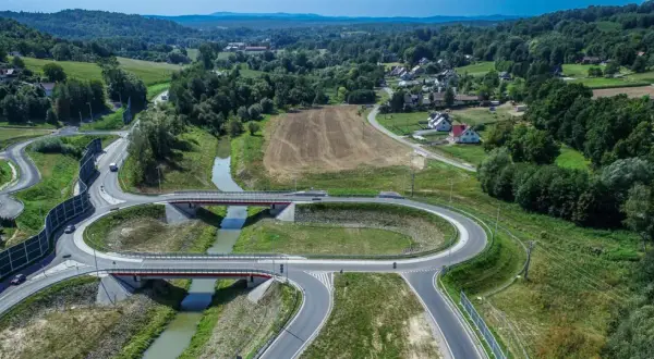Nowoczesny węzeł drogowy na drodze krajowej 75, z mostami nad rzeką, otoczony zielonymi wzgórzami i domami.