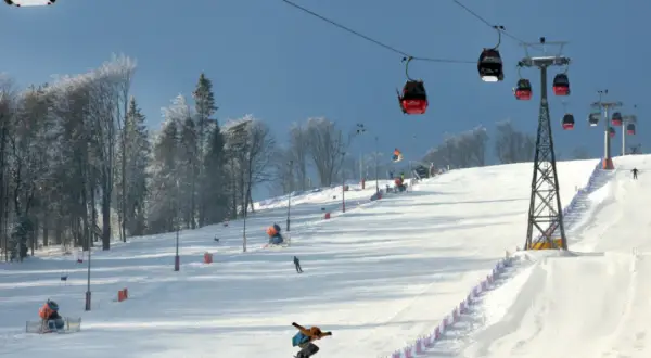 Kolejka gondolowa nad ośnieżonym stokiem narciarskim, na którym snowboardzista wykonuje skok.