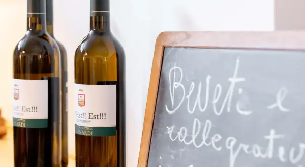 Bottiglie di vino "Est!! Est!!!" in primo piano, con una lavagna che invita a "Bevete e rallegratevi".