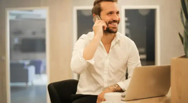 Uśmiechnięty mężczyzna w białej koszuli rozmawia przez telefon, siedząc przy biurku z laptopem. Może właśnie dowiaduje się, które banki dzwonią do pracodawcy.
