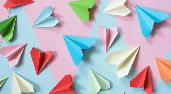 Kolorowe papierowe samolociki, jak się robi origami. Różne odcienie papieru ułożone na tle geometrycznych kształtów.
