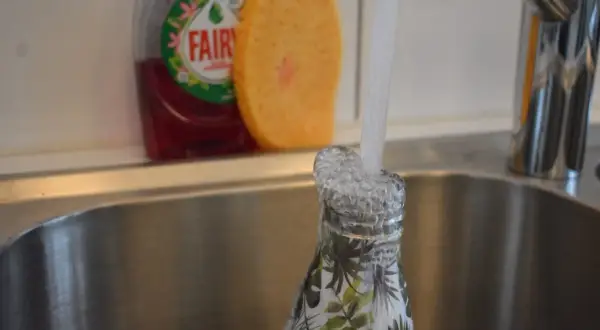 Eine Nalgene-Flasche mit Blattmuster wird in der Spüle unter fließendem Wasser gespült. Spülmittel und Schwamm sind im Hintergrund sichtbar.
