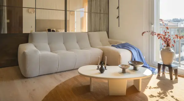 Divano chiaro e tavolino bianco su un tappeto di colore marrone. Sul tavolino, vasi e candele. Sul divano, una coperta blu.