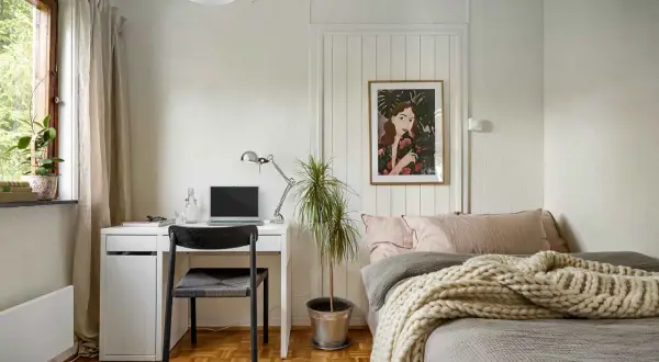Gemütliches homeoffice im schlafzimmer mit Schreibtisch, Laptop und Bett. Ein Bild mit einer Frau und Pflanzen ziert die Wand.