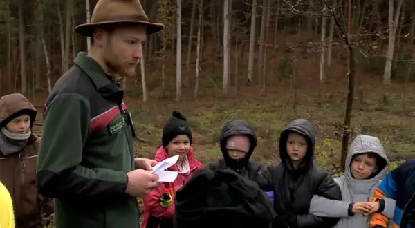 Ein Förster mit braunem Hut erklärt Kindern im Wald etwas. Sie sind von Hütten aus hierher gekommen, um mehr über die Natur zu lernen.